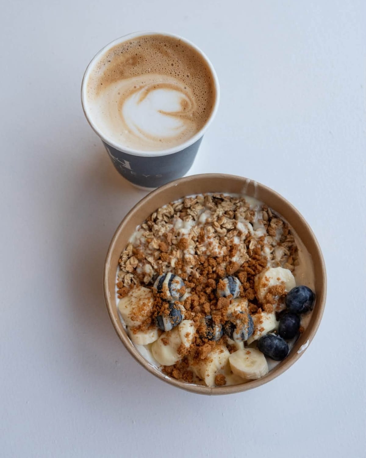 A latte in a Hedleys cup next to a yoghurt bowl with granola, banana and blueberries